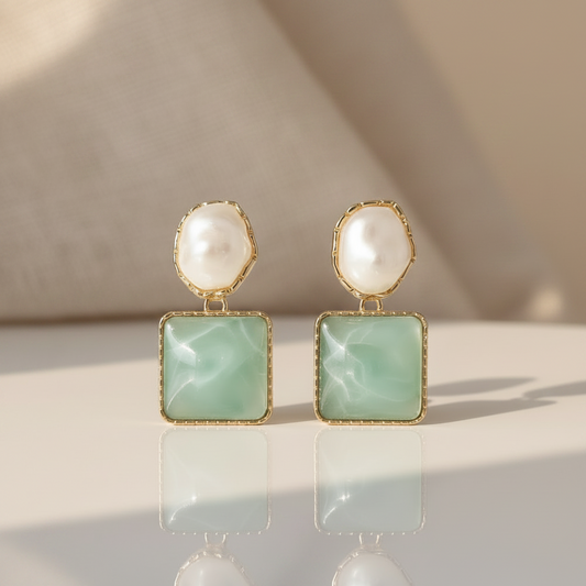 Pair of earrings with gold frames, white pearls, and green square stones on a light gray background.
